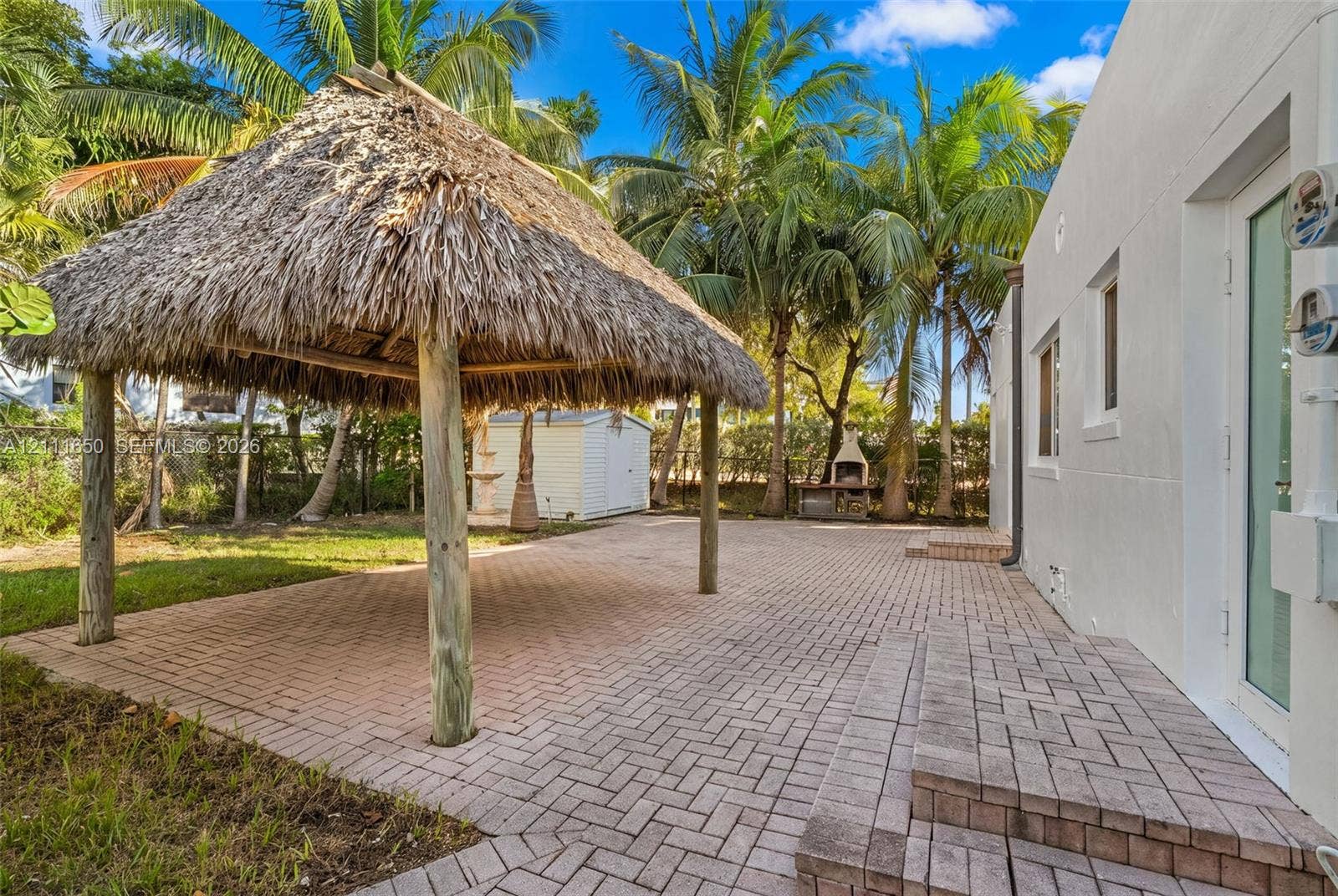 Backyard featuring a thatched-roof tiki hut, extensive brick paver patio, stone-finish outdoor grill, and mature palm trees