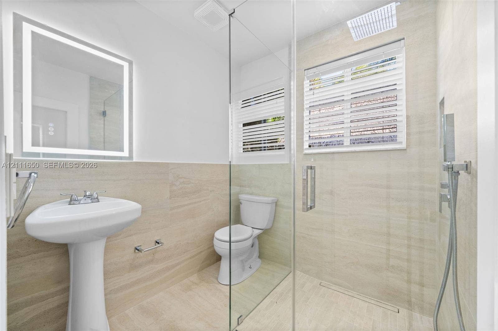 Bathroom featuring stone-finish wall tiling, a pedestal sink, a walk-in shower with a glass enclosure and handheld faucet, and an illuminated wall-mounted mirror