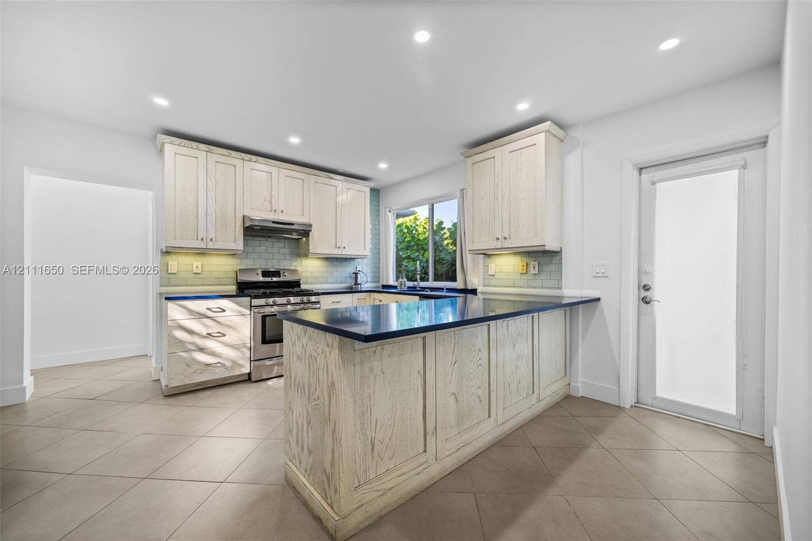 Kitchen featuring light-toned wood cabinetry, dark solid-surface countertops, a subway tile backsplash, recessed lighting, and tile flooring
