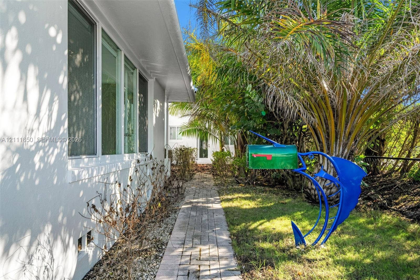 Exterior side profile featuring smooth-finish white stucco walls, multi-pane windows, and a paved brick walkway bordered by mature palms and foliage