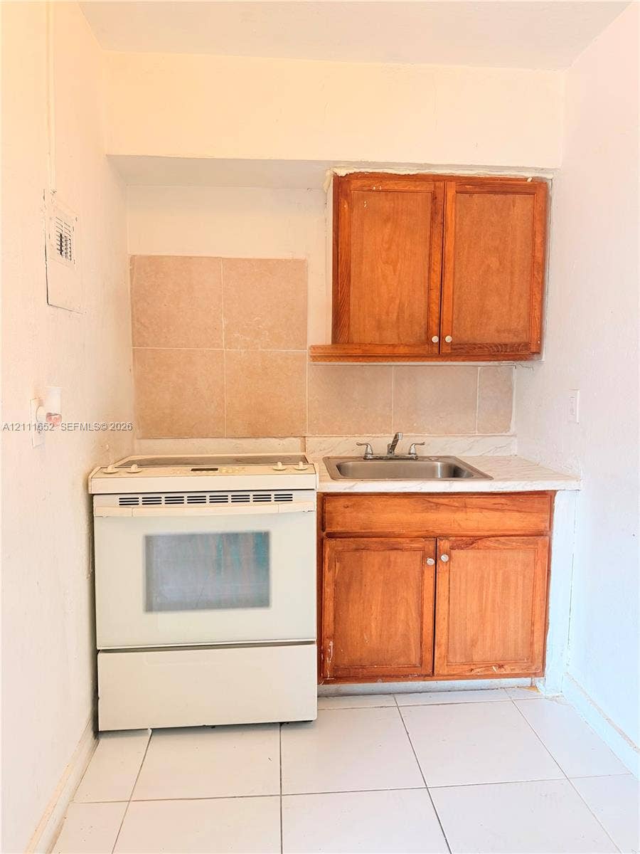 Kitchenette featuring white porcelain floor tiles, wood-finish cabinetry, a single-basin stainless steel sink, a white electric range, and a tiled backsplash