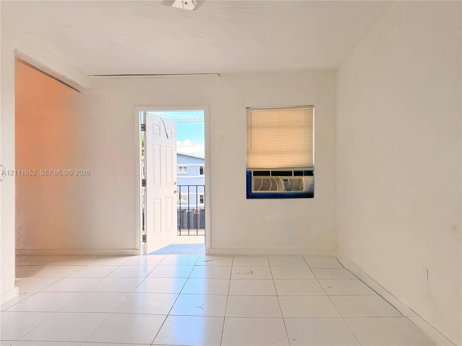 Interior space featuring white ceramic tile flooring, a window-integrated air conditioning unit, and a doorway opening to a metal-railed exterior balcony