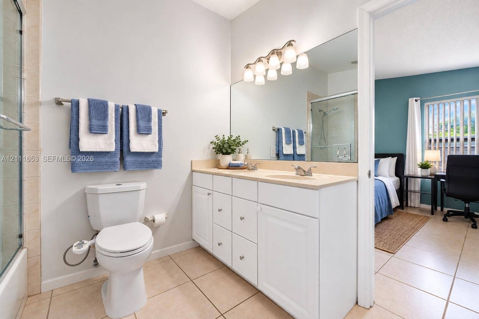 1042 NE 38th Street, Oakland Park, FL 33334 Primary Bathroom featuring white cabinetry, solid-surface vanity countertop with dual integrated sinks, full-width wall mirror, and tile flooring