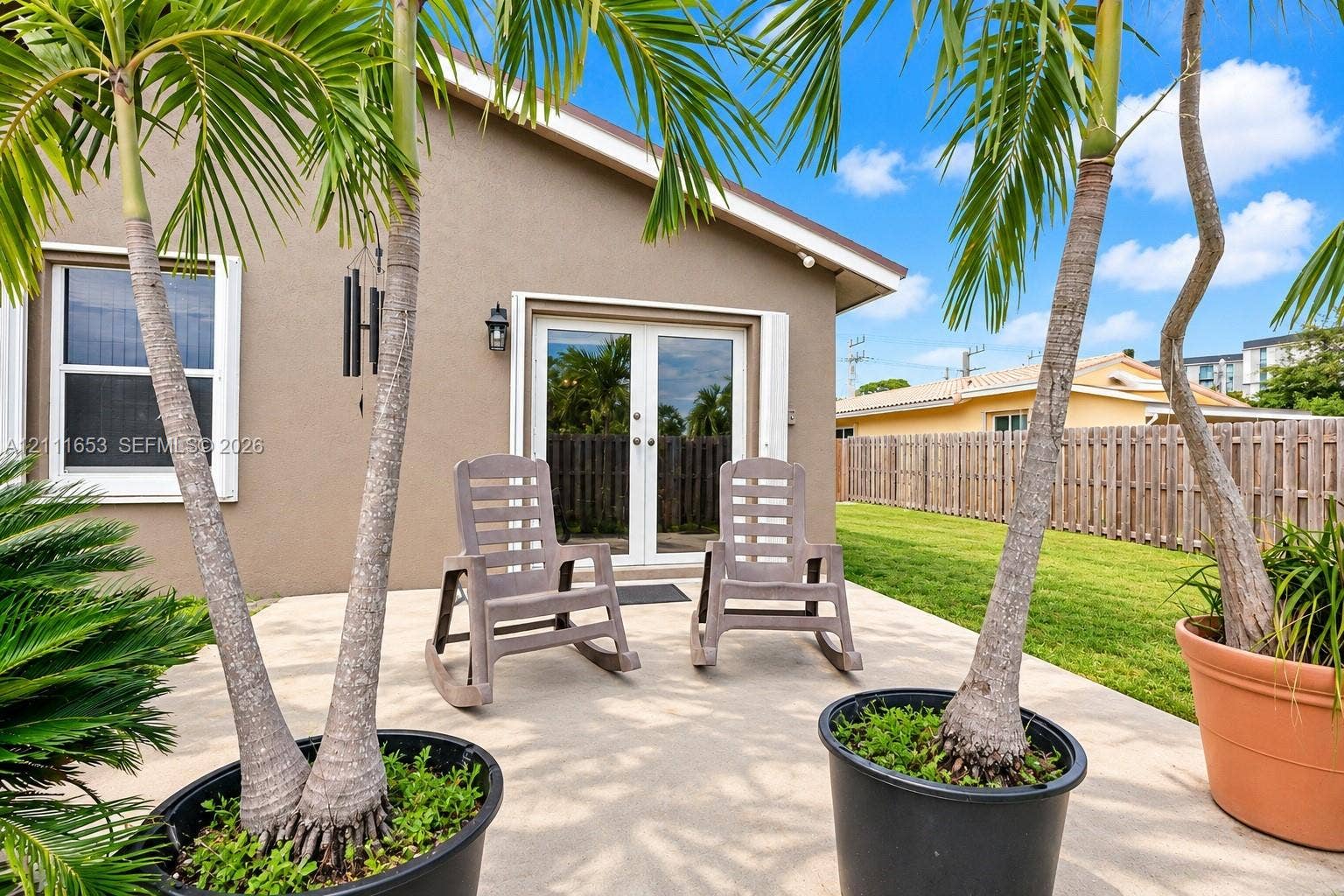 1042 NE 38th Street, Oakland Park, FL 33334 Concrete patio featuring a set of exterior French doors, an attached exterior wall-mounted light fixture, and an adjoining lawn enclosed by a wooden fence