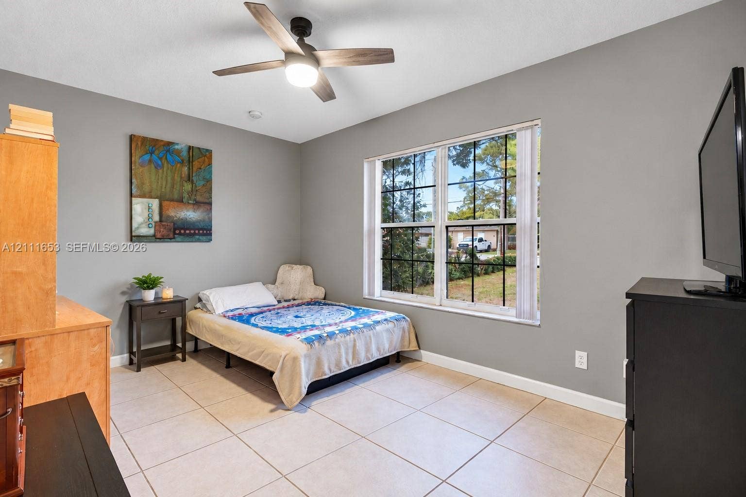 1042 NE 38th Street, Oakland Park, FL 33334 Bedroom 3 Neutral-toned walls featuring large-format ceramic tile flooring, a multi-pane window with grid detailing, and a modern ceiling fan with integrated lighting