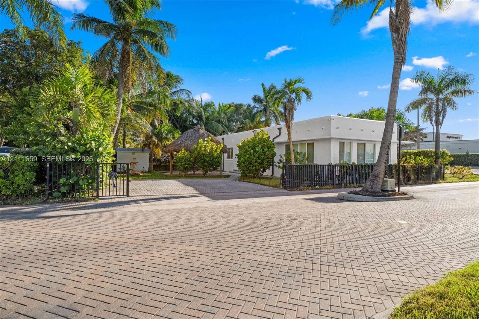 338 Taylor Street, #2, Hollywood, Hollywood, FL 33019 Exterior featuring a white flat-roof facade, a herringbone-patterned brick paver driveway, an outdoor thatched-roof structure, and perimeter metal fencing