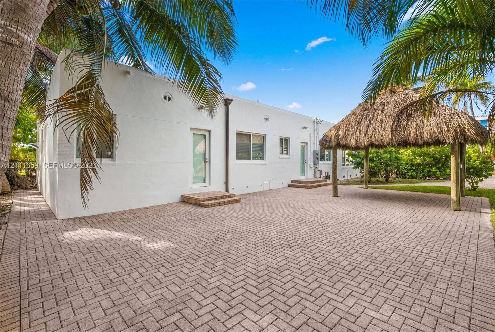 338 Taylor Street, #2, Hollywood, Hollywood, FL 33019 Rear exterior featuring a herringbone-pattern paver patio, thatched-roof tiki hut, white stucco facade, and mature palm trees