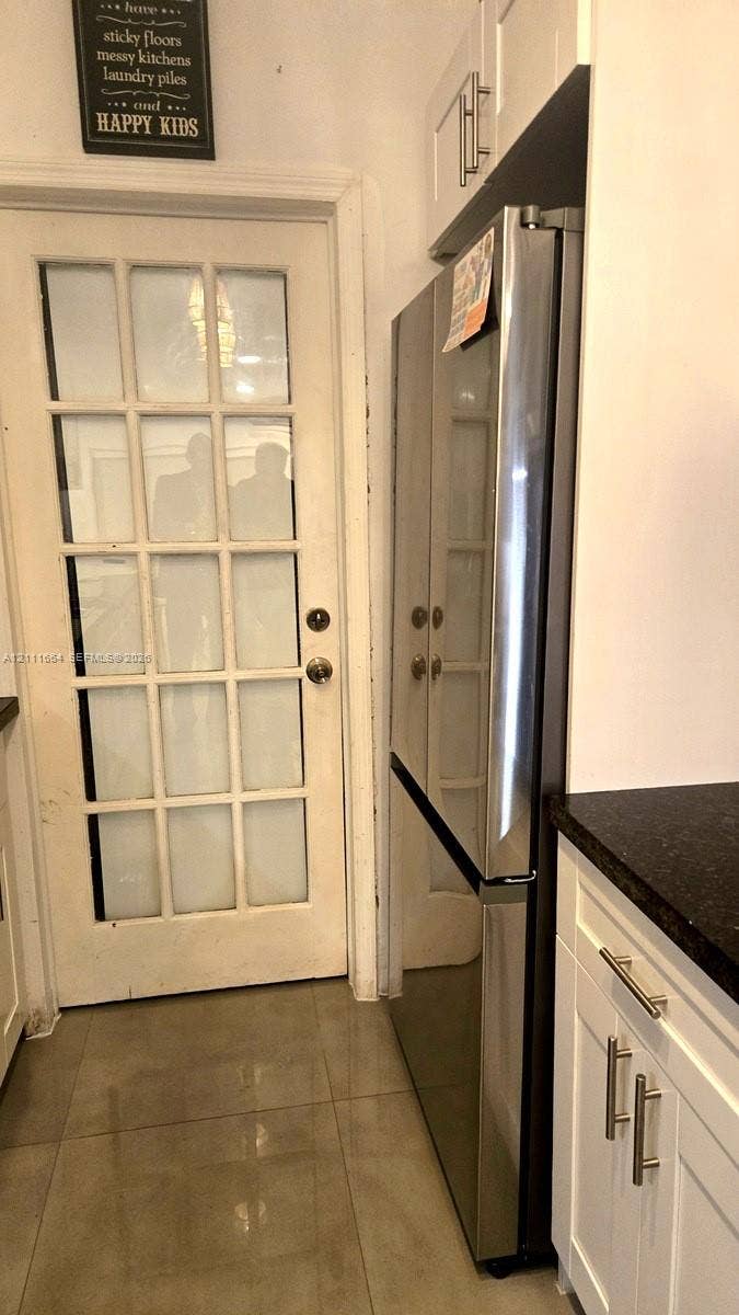 Stainless steel refrigerator, shaker-style white cabinetry, dark stone-finish countertop, and white-framed glass-panel door, set against tile flooring