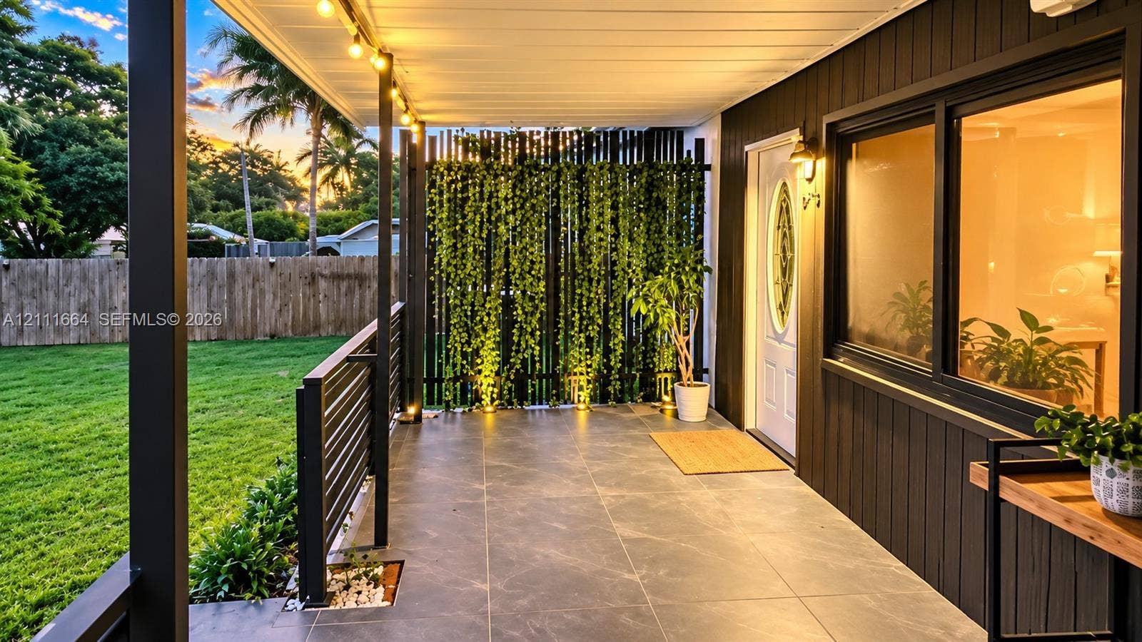 Covered patio featuring large-format stone-finish tile flooring, horizontal slat privacy screening, a black metal railing, and exterior wall-mounted lighting