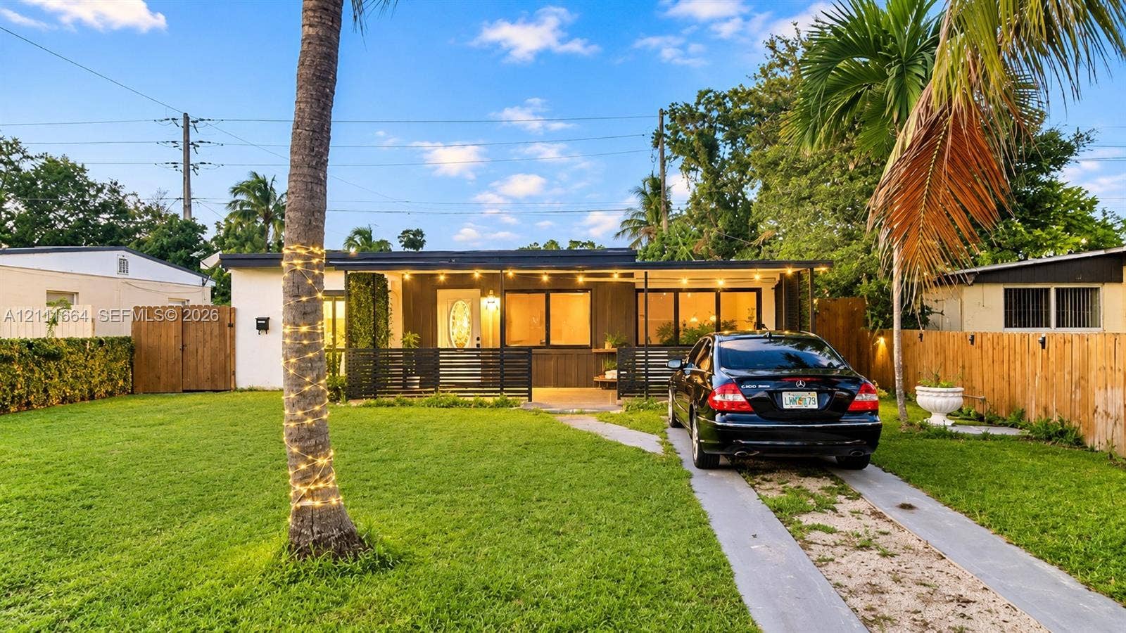 Single-story exterior featuring a covered front porch, horizontal wood-panel siding, mature palm trees, and a concrete-strip driveway