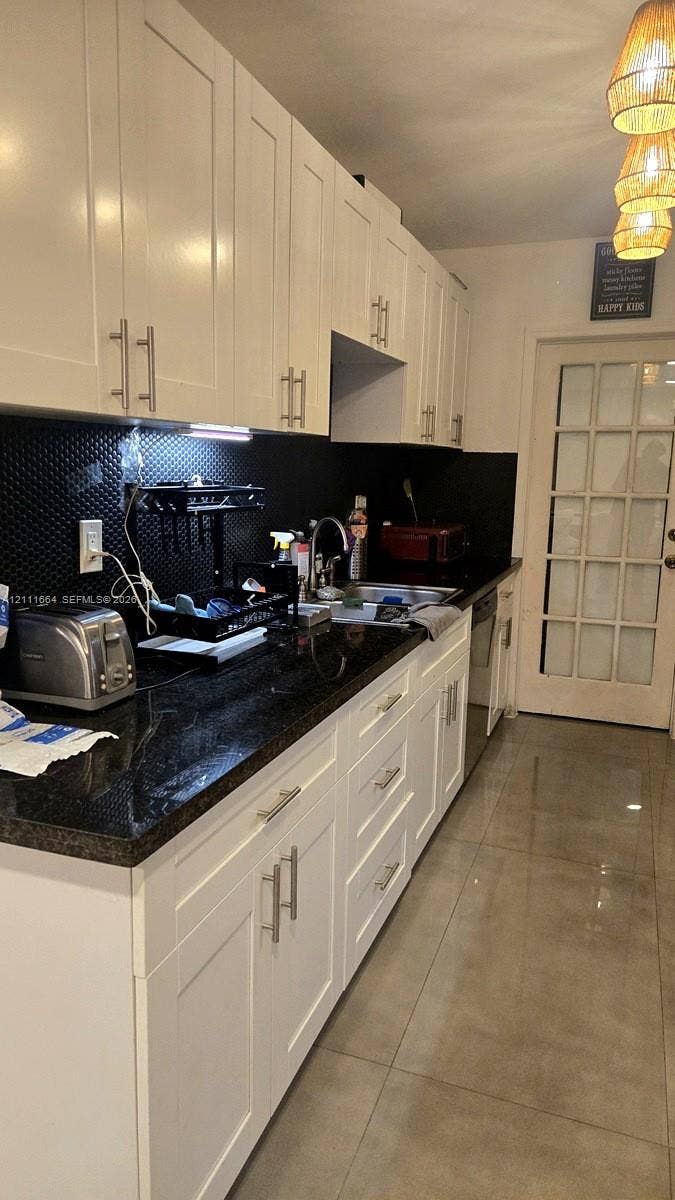 Kitchen featuring white shaker-style cabinetry, dark stone countertops, a textured black backsplash, large-format tile flooring, and a glass-paneled door