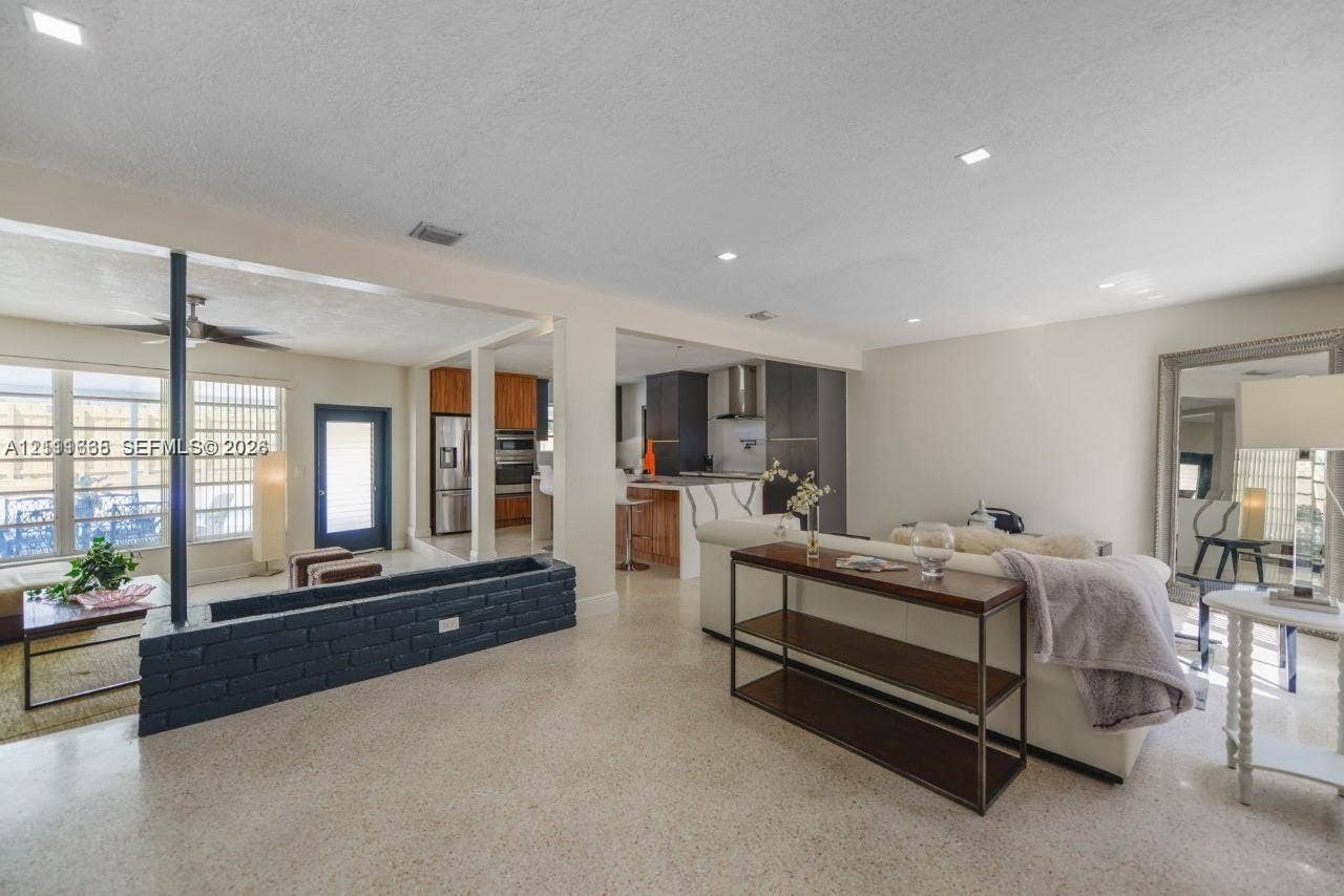 Open-concept interior featuring light-toned stone flooring, a painted brick structural partition, recessed lighting, and a kitchen equipped with dark cabinetry and a central island