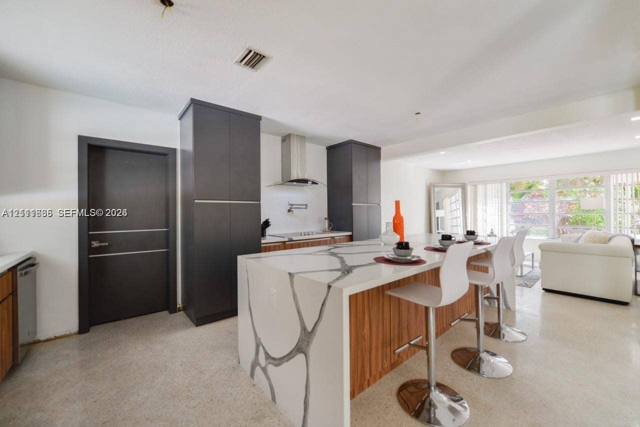 Kitchen featuring a waterfall-edge island with stone-finish countertops and wood-finish cabinetry, floor-to-ceiling dark charcoal pantry units, and a stainless steel range hood