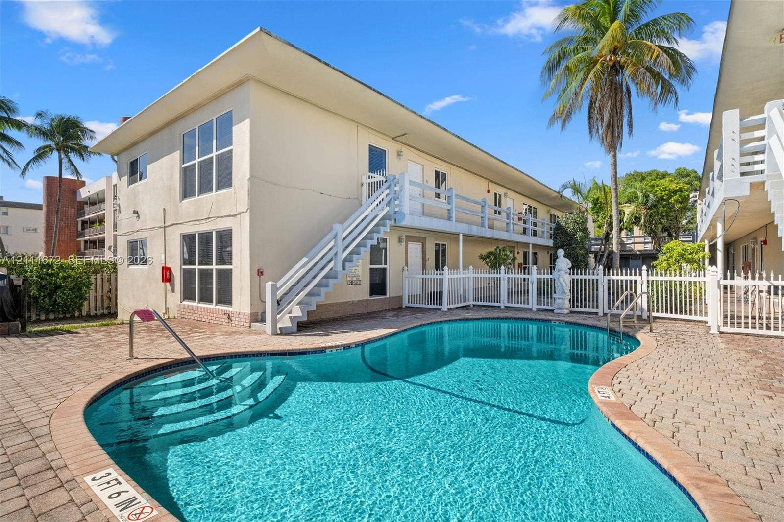 In-ground swimming pool with stone-paver decking, white picket fencing, and two-story building exterior featuring exterior staircases