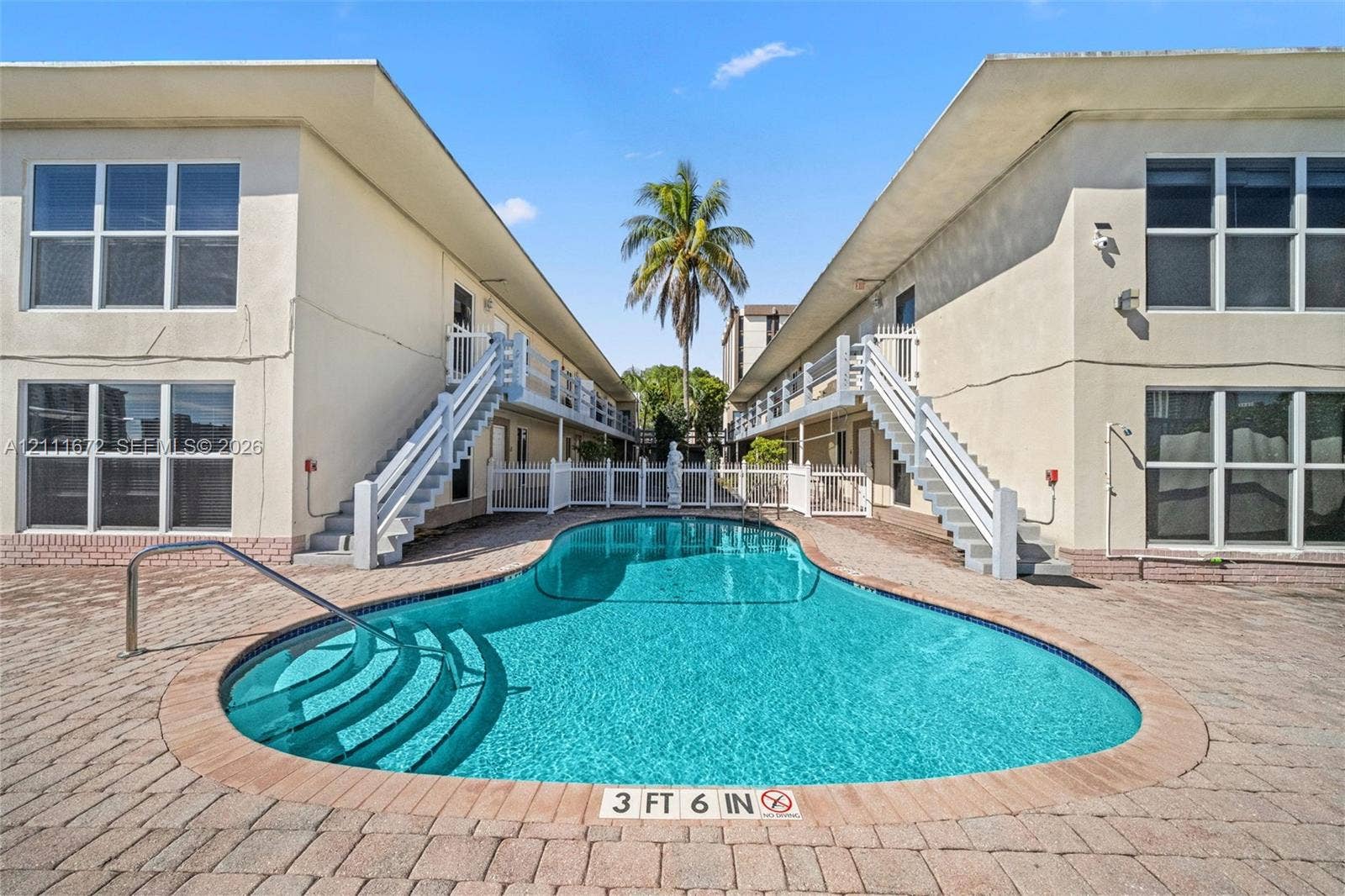 In-ground swimming pool featuring curved entry steps, surrounded by a brick paver deck, dual staircases with white railings, and multi-pane window configurations