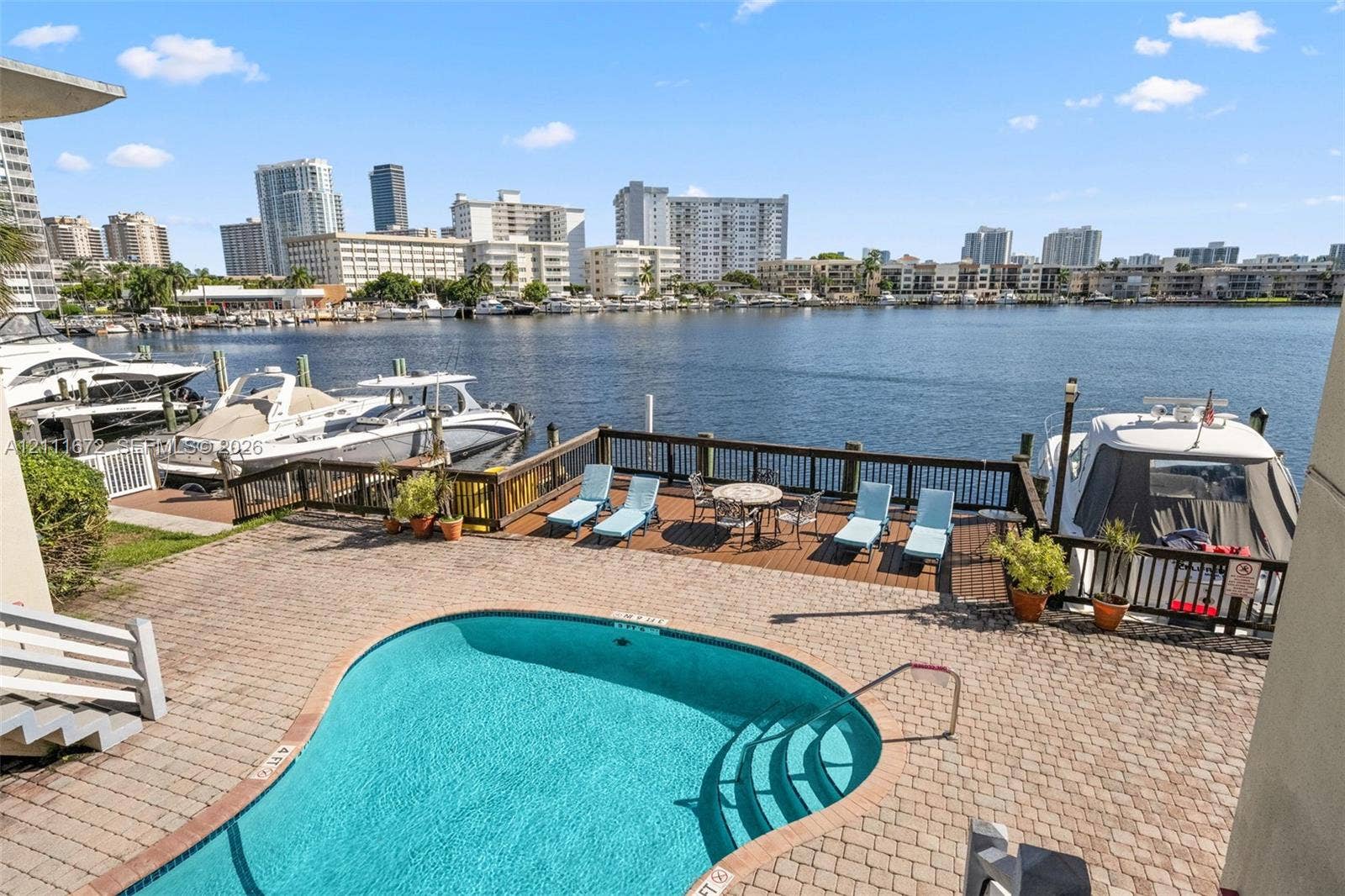 Waterfront terrace featuring a curvilinear swimming pool, paver hardscaping, and a private wooden dock overlooking a marina