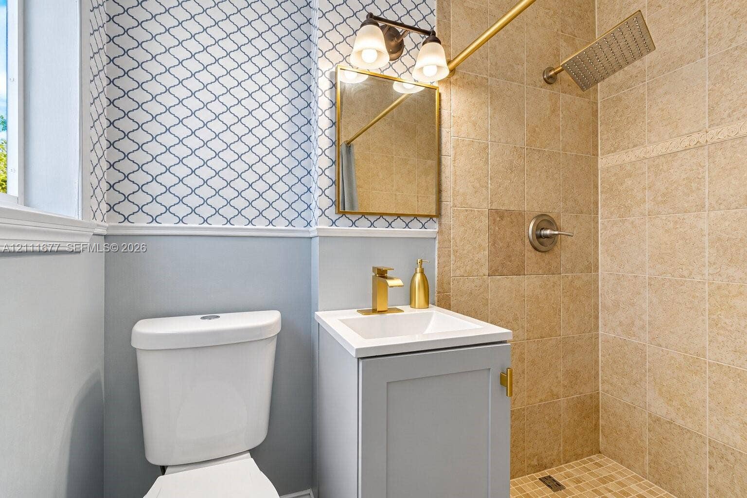 1921 Acapulco Drive, Miramar, FL 33023 Bathroom featuring a vanity with a white rectangular basin, brass-finish faucet, framed wall-mounted mirror, two-light fixture, and a walk-in shower with square tile surround and rainfall showerhead