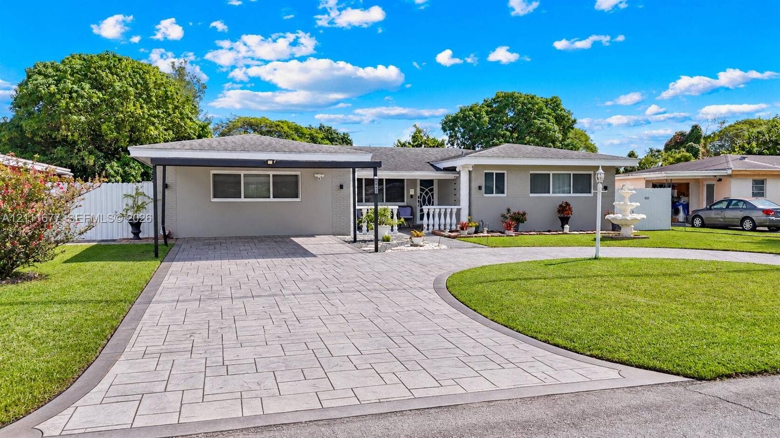 1921 Acapulco Drive, Miramar, FL 33023 Single-story exterior featuring a paver driveway, covered carport, manicured lawn, tiered water fountain, and white balustrade railing