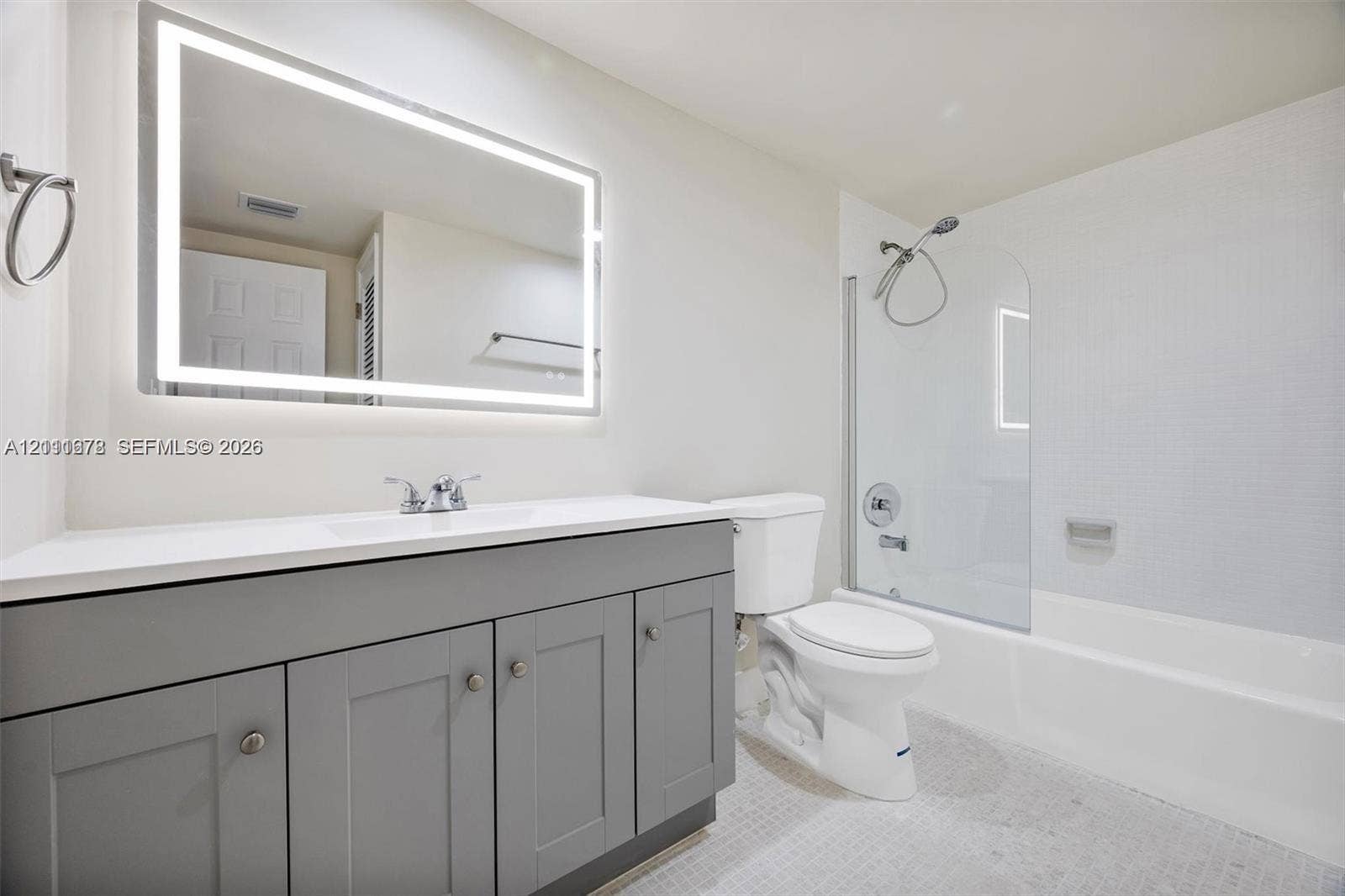 Bathroom featuring a grey shaker-style vanity with a solid surface countertop, an illuminated rectangular wall mirror, a glass shower partition, and a built-in bathtub with a wall-mounted shower head