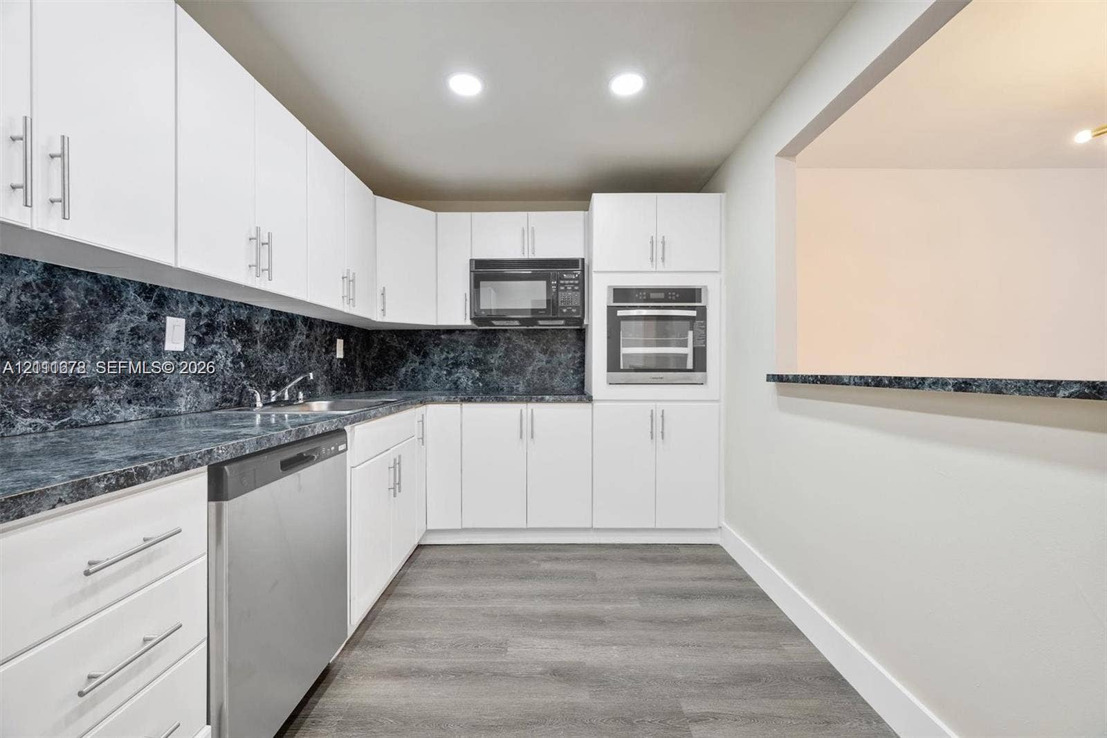 Kitchen featuring white shaker-style cabinetry, dark stone-patterned countertops, integrated stainless steel oven and microwave, stainless steel dishwasher, and wood-finish flooring