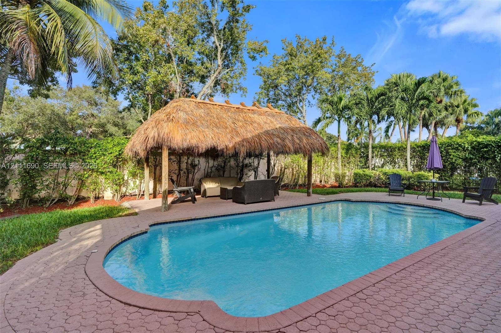 In-ground pool surrounded by paver patio, featuring a built-in thatched-roof tiki hut and privacy hedging