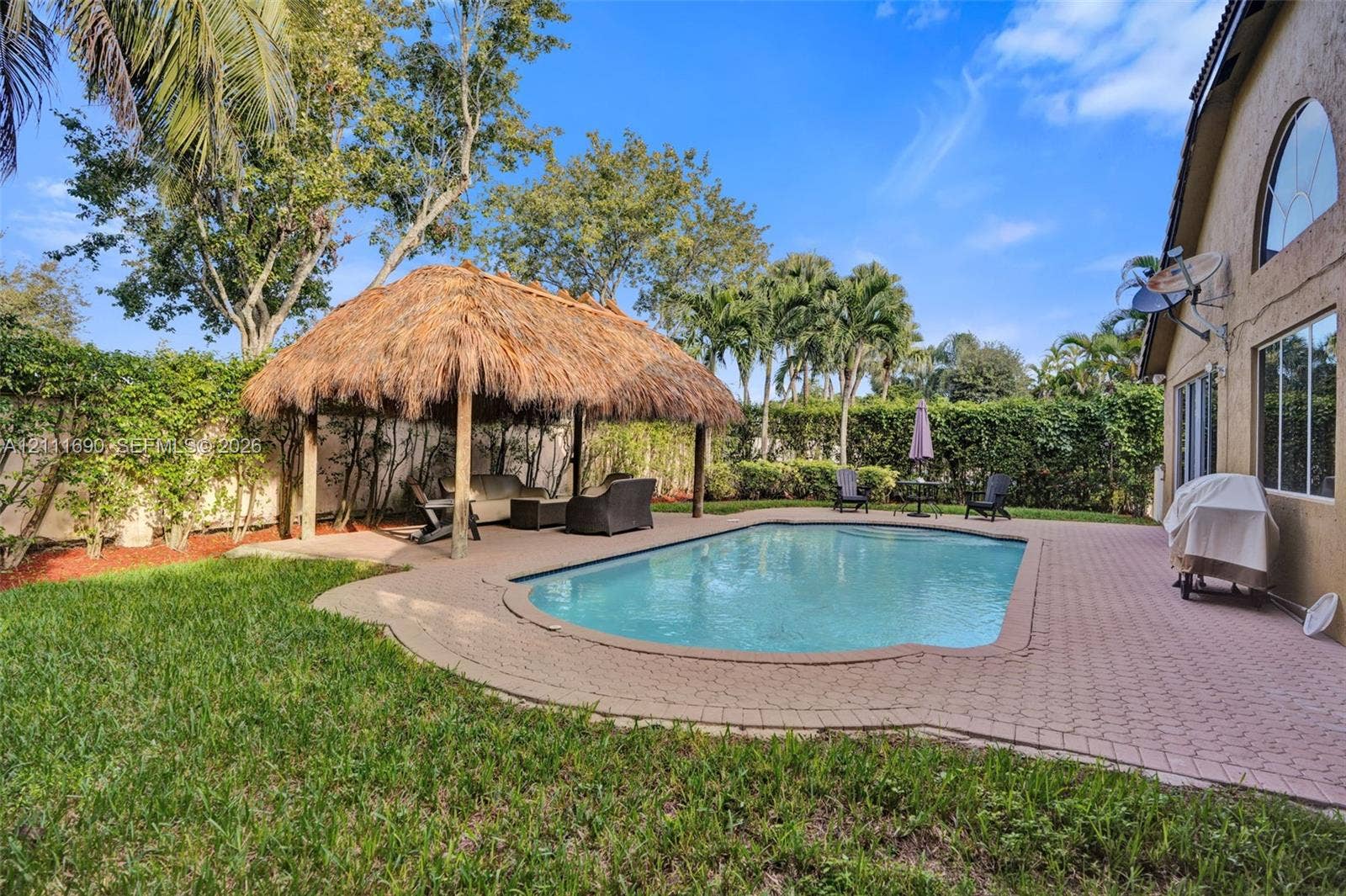 In-ground swimming pool with paver decking, thatched-roof tiki hut, and privacy-hedged backyard