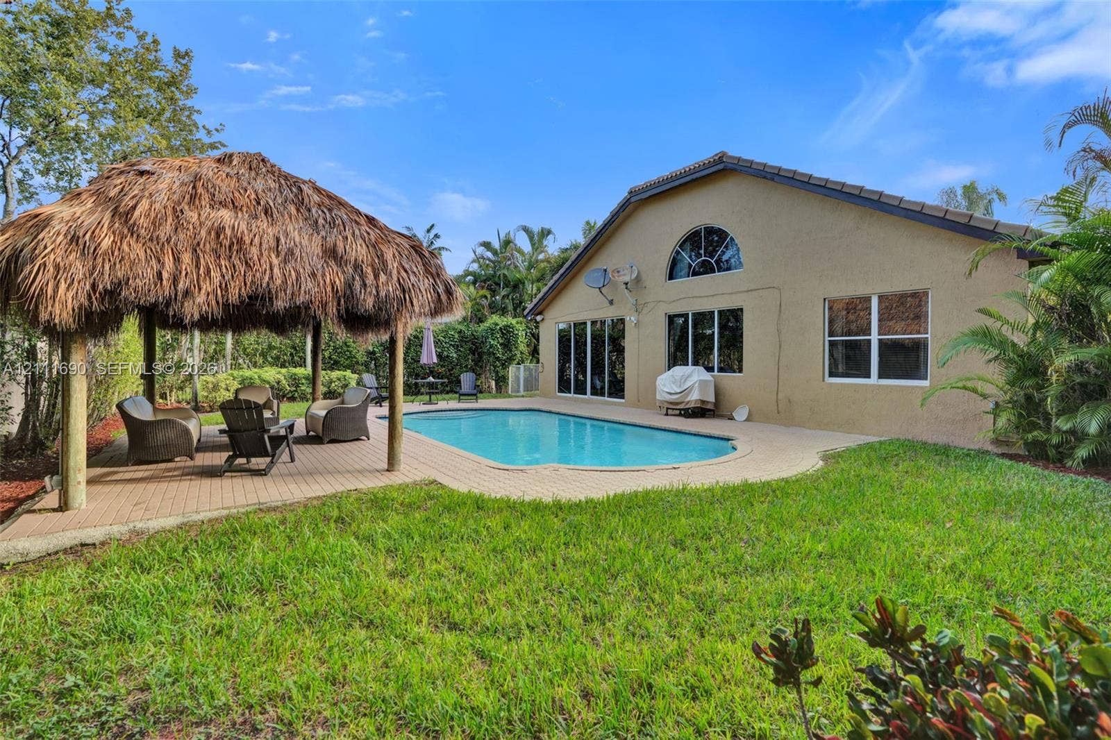 Stucco-exterior residence featuring an in-ground swimming pool, a thatched-roof tiki hut, a paved patio, and a manicured lawn