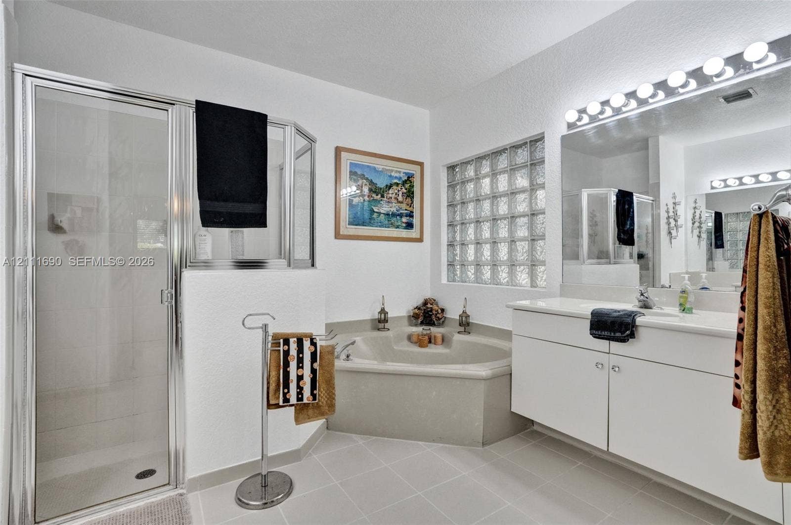 Bathroom featuring a walk-in shower with a glass enclosure, a corner soaking tub, a white vanity with integrated basin, glass block accent window, and a multi-bulb vanity light fixture