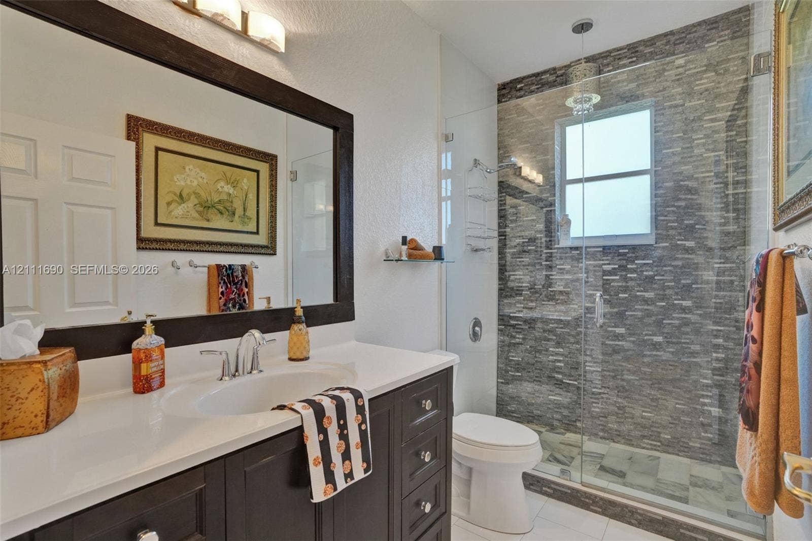 Dark wood vanity with a solid surface countertop, glass-enclosed shower featuring stone-finish wall tile, and a framed mirror with integrated lighting