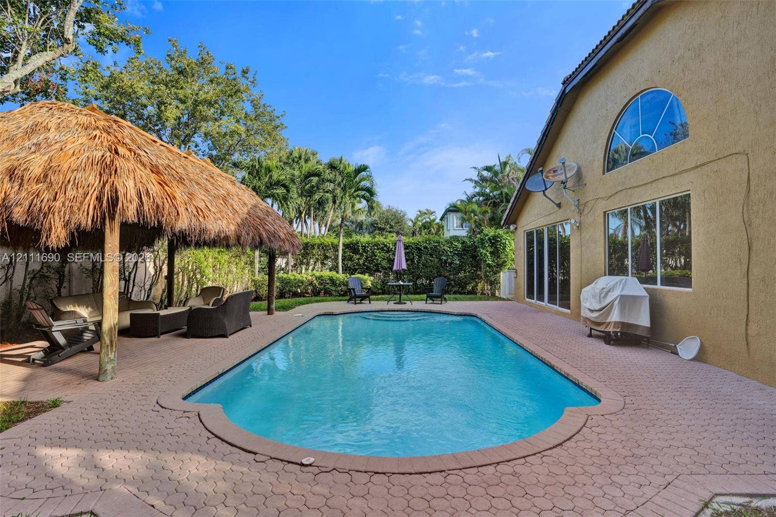 Swimming pool with concrete paver decking, thatched-roof cabana, and mature perimeter landscaping