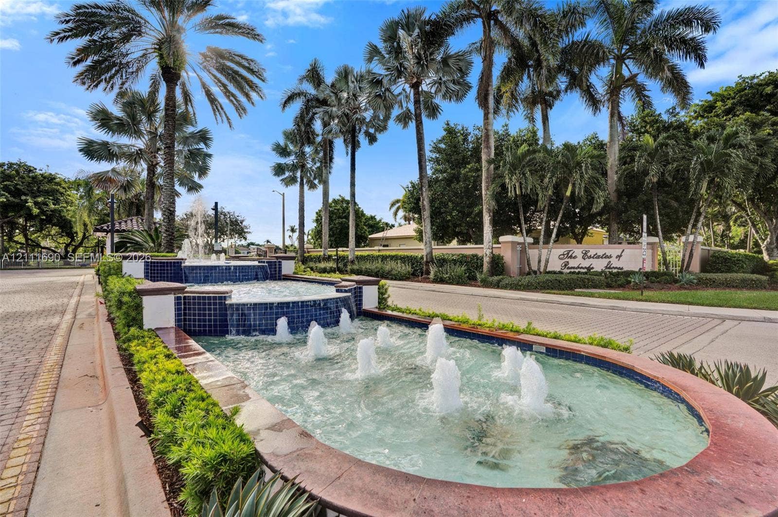 Tiered water fountain featuring blue tile accents, surrounded by manicured landscaping, mature palm trees, and brick-paved thoroughfares