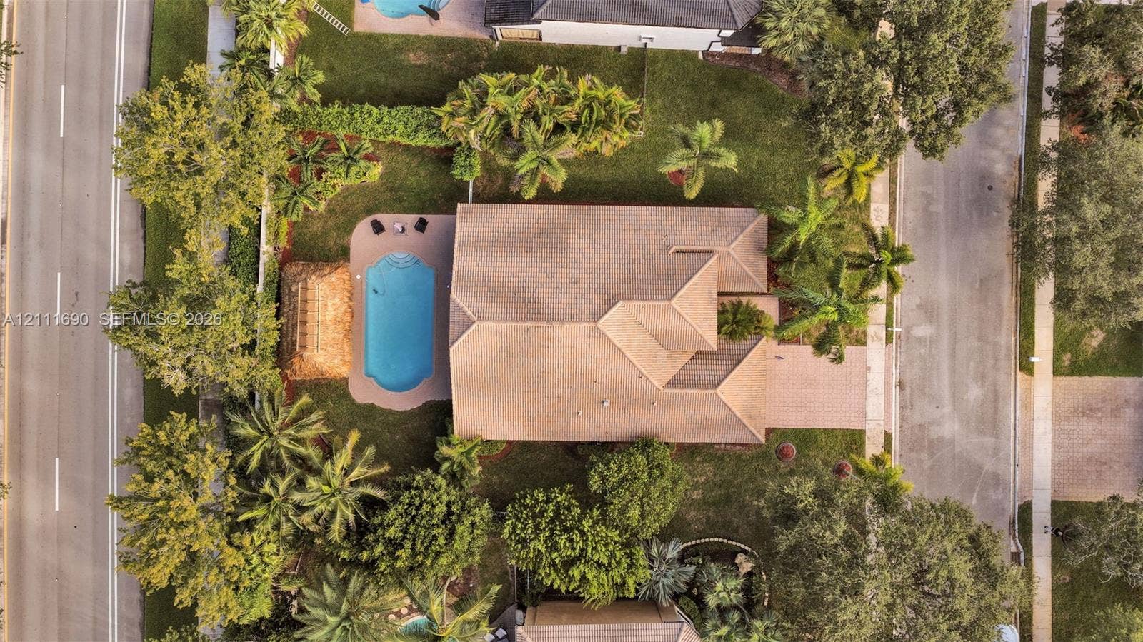 Aerial perspective of a property featuring a swimming pool, paved patio, lush landscaping with mature palms, and a multi-pitched roof