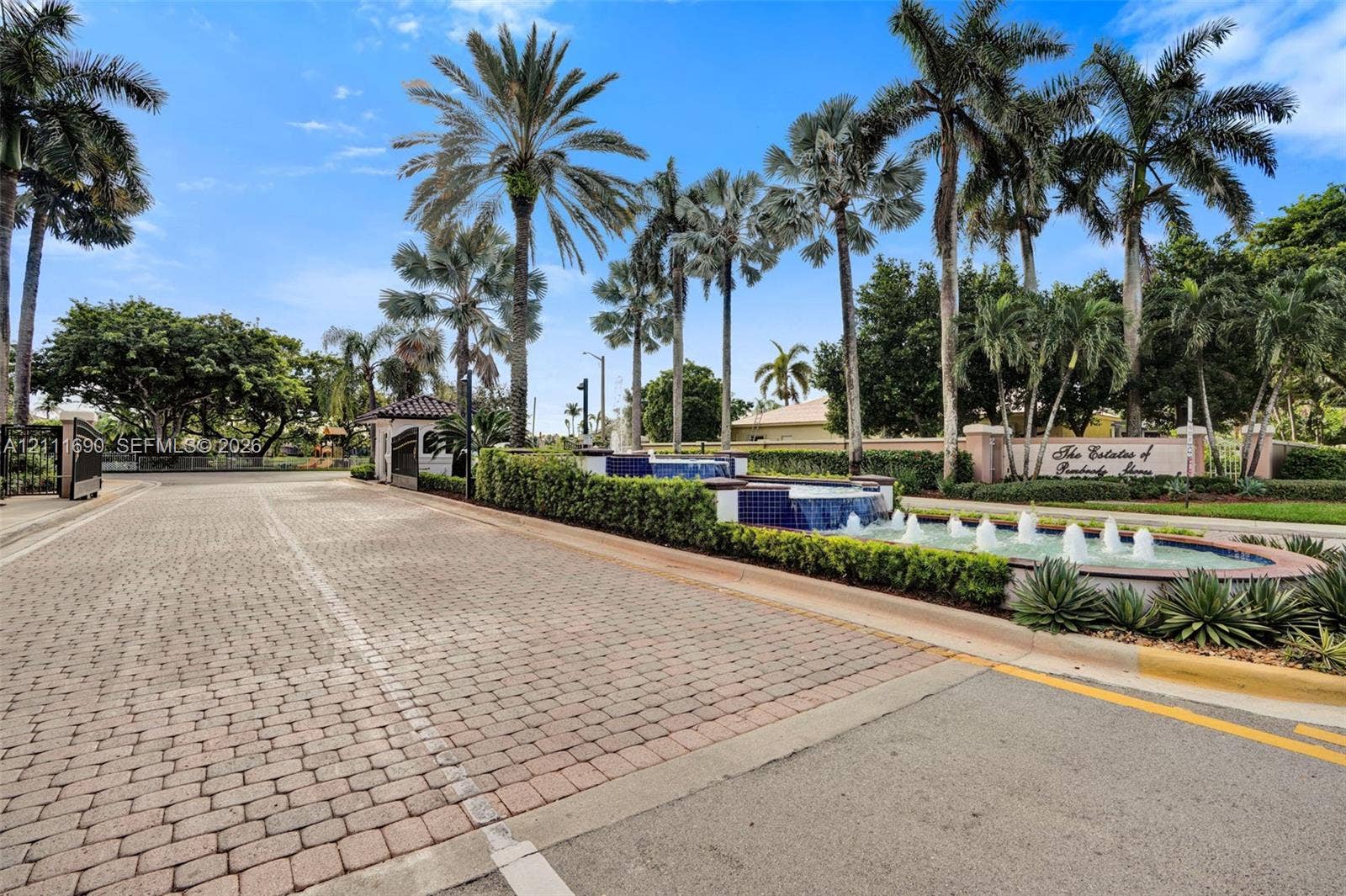 Gated community entrance featuring a paver brick roadway, multi-tiered stone fountain, and mature palm landscaping