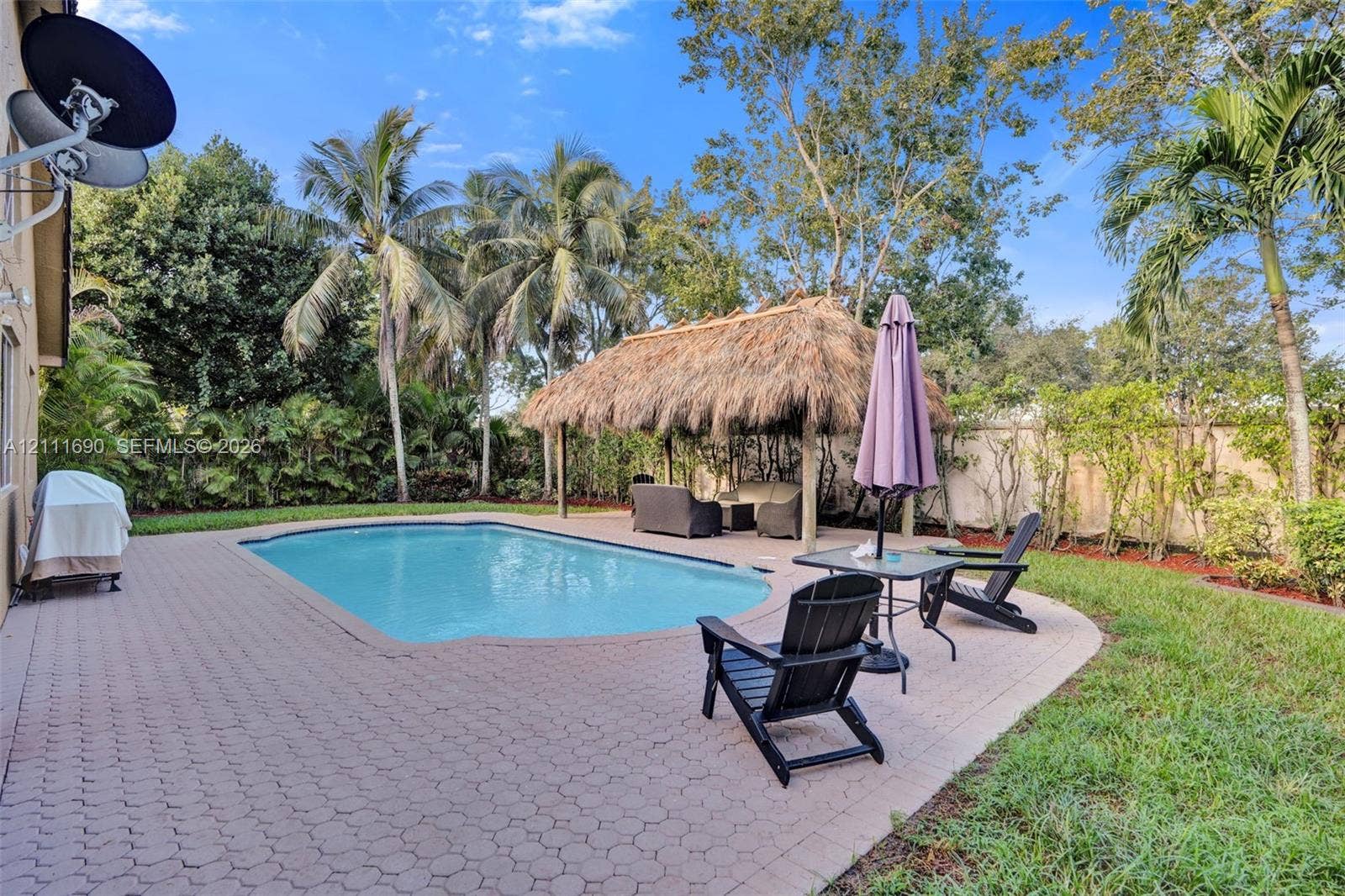 Outdoor space featuring a concrete paver patio, in-ground swimming pool, and a thatched-roof tiki hut, surrounded by mature palm trees and lush landscaping