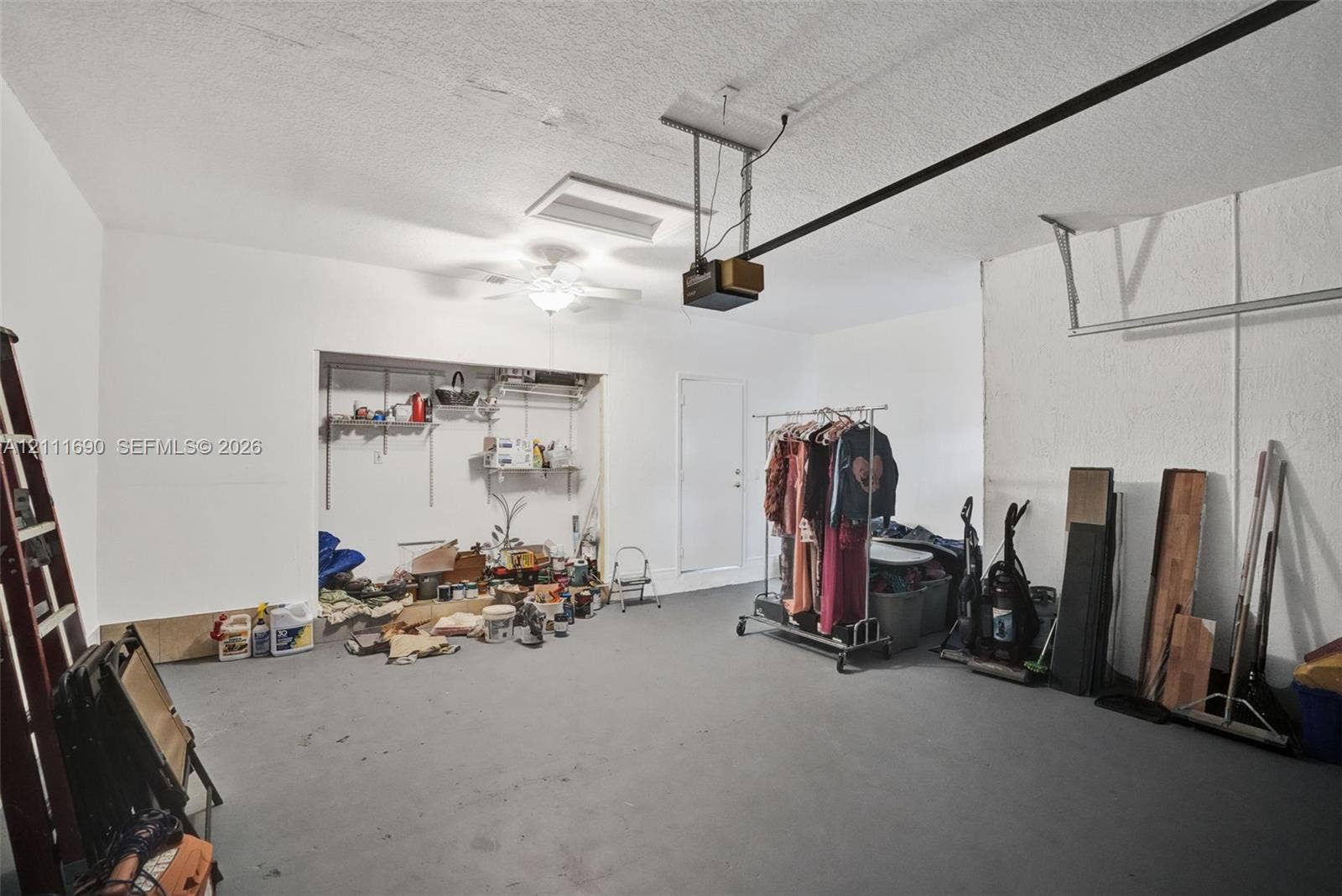 Garage interior featuring a painted concrete floor, ceiling-mounted automatic door opener, ceiling fan with light fixture, and recessed storage alcove
