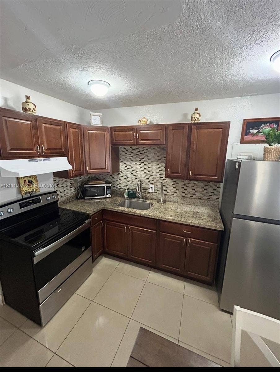 Kitchen featuring wood cabinetry, stone-finish countertops, tile backsplash, stainless steel appliances, and tile flooring