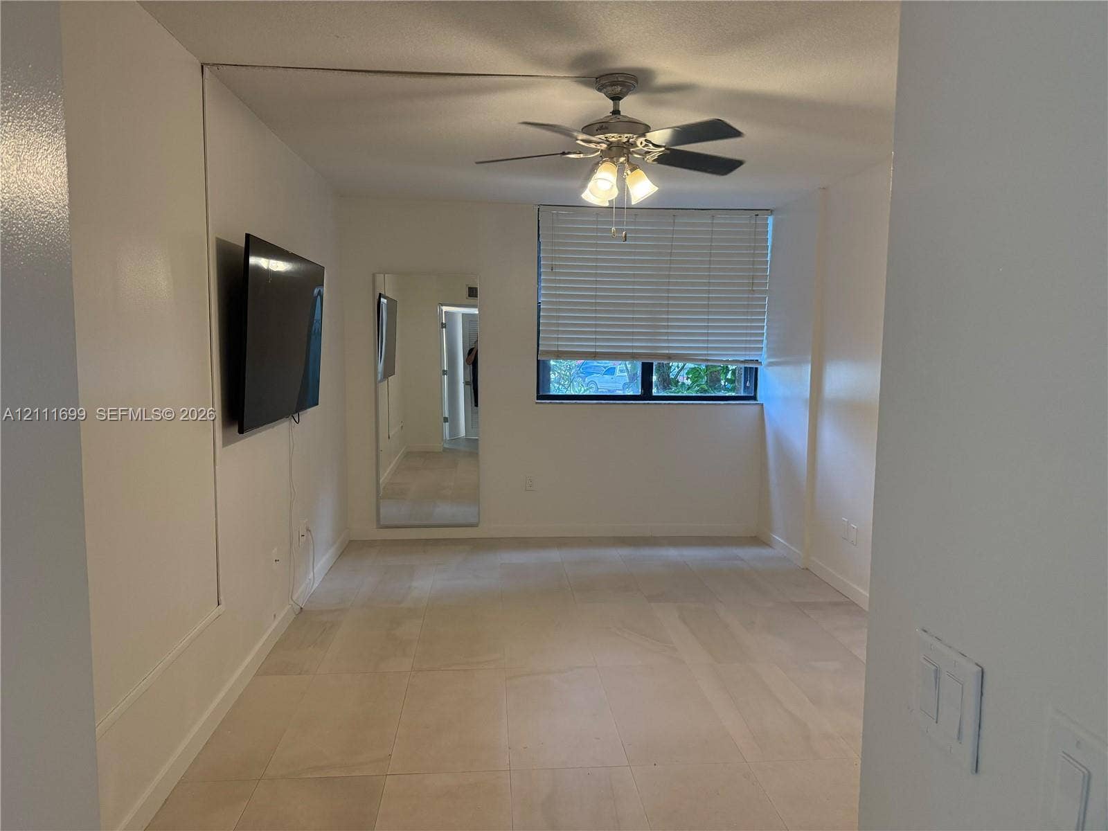 Neutral-toned large format tile flooring, ceiling fan with integrated light fixture, full-height wall mirror, and window with horizontal blinds