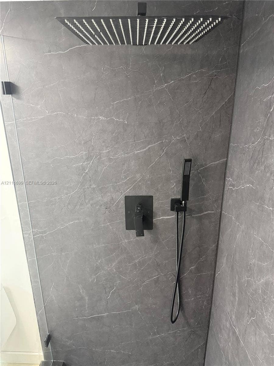 Shower enclosure featuring grey stone-finish tile surround, oversized square rainfall showerhead, matte black handheld spray, and coordinated wall-mounted control hardware