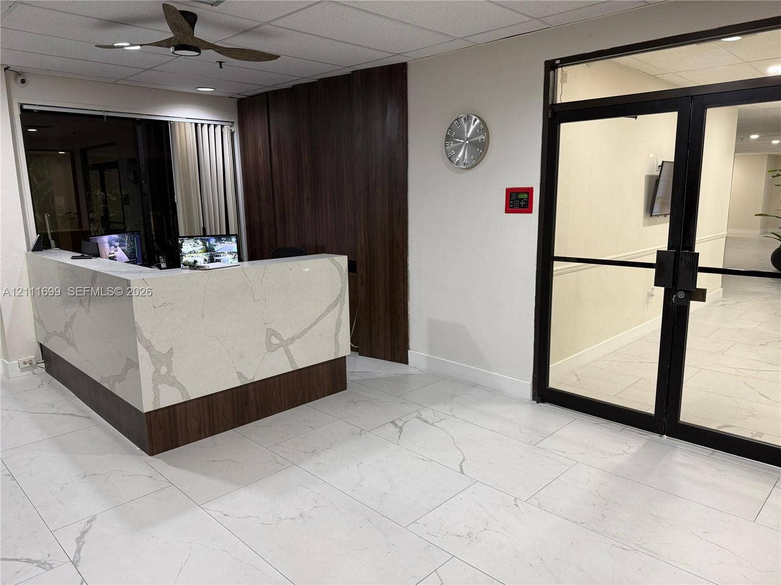 Reception area featuring a stone-topped built-in desk with wood-finish paneling, large-format tile flooring, a dark-finish ceiling fan, and double glass doors with black framing