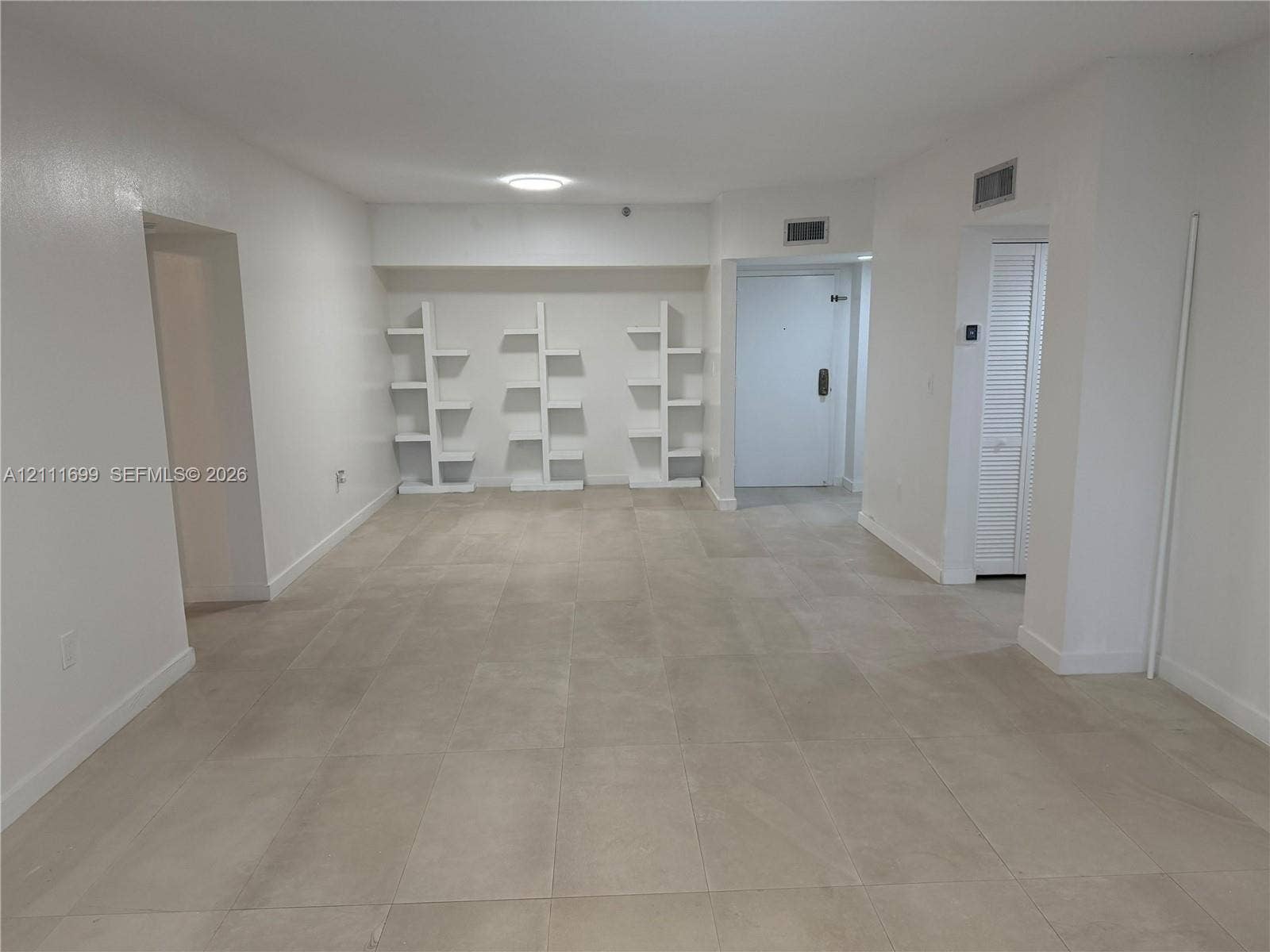 Neutral-toned floor tiles, three built-in white tiered shelving units, and a closet with louvered doors