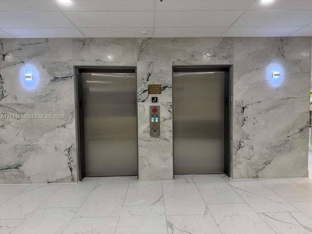 Dual stainless steel elevator doors featuring floor-to-ceiling stone-clad surround, large-format tile flooring, and wall-mounted light fixtures