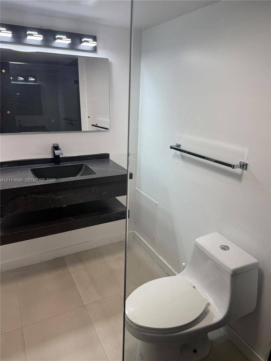 Floating vanity with a dark-toned stone surface and integrated basin, matte black faucet, wall-mounted rectangular mirror with integrated lighting, and one-piece white toilet