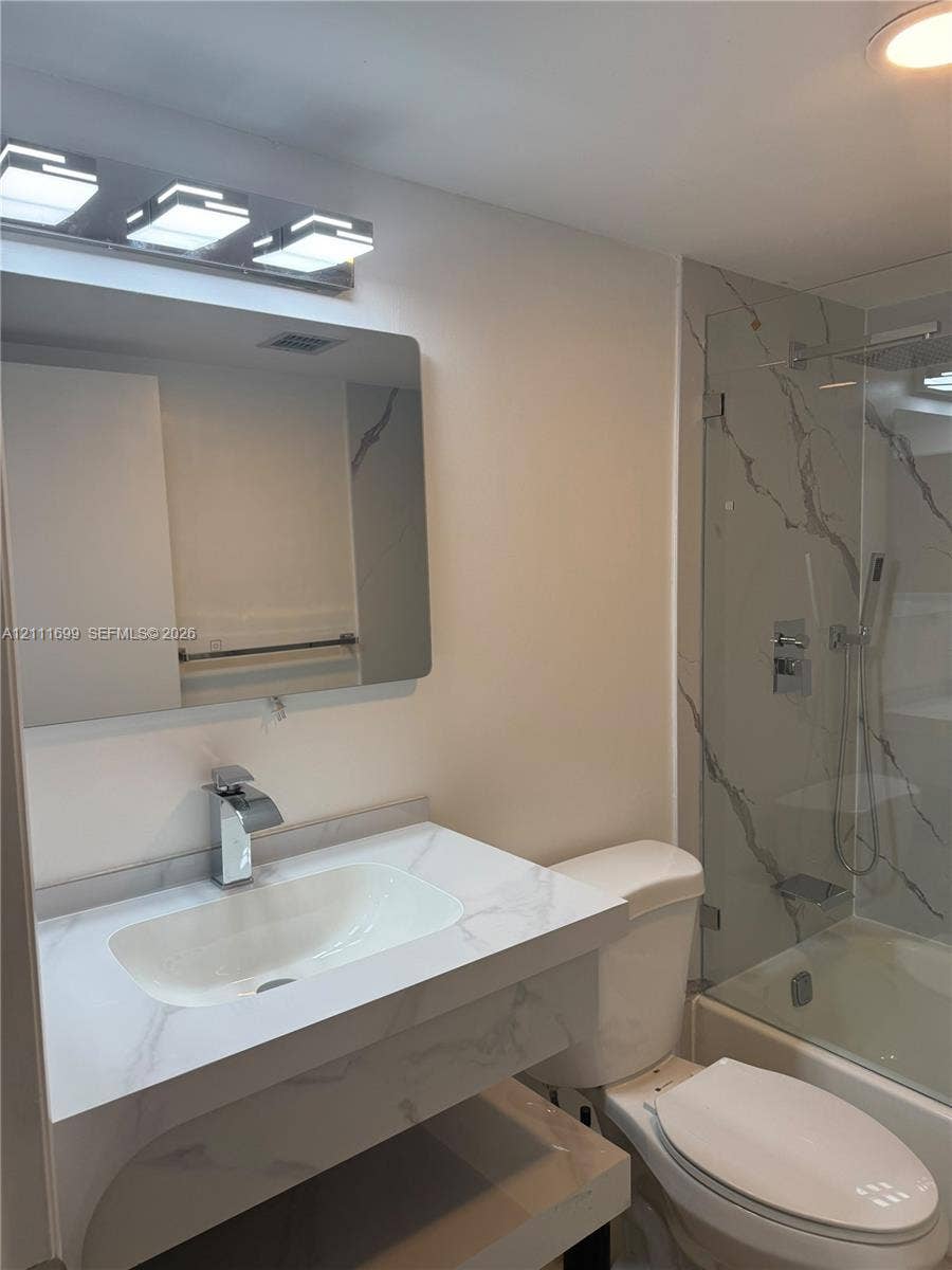 Bathroom featuring a vanity with stone-finish countertop and integrated sink, modern rectangular chrome faucet, wall-mounted mirror, tiered chrome light fixture, and a glass-enclosed shower with floor-to-ceiling veined stone-finish tile surround