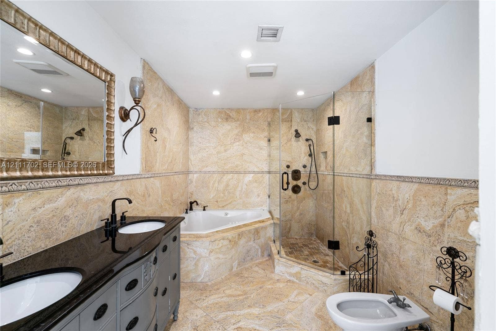 Dual-sink vanity with dark stone countertop, stone wall and floor tile, jetted soaking tub, and glass-enclosed shower with body spray system