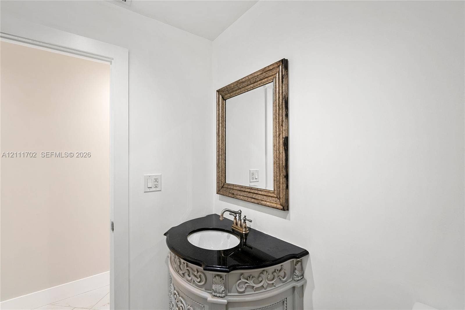Ornate light-grey vanity with decorative carved detailing, black stone countertop, integrated undermount sink, brushed-metal faucet, and wall-mounted framed mirror