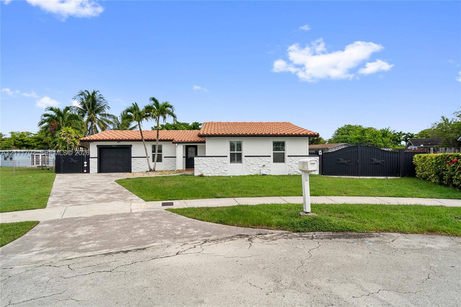 Single-story exterior featuring white stucco walls with textured stone facade accents, a multi-gabled clay tile roof, an attached garage, and black ornamental metal fencing
