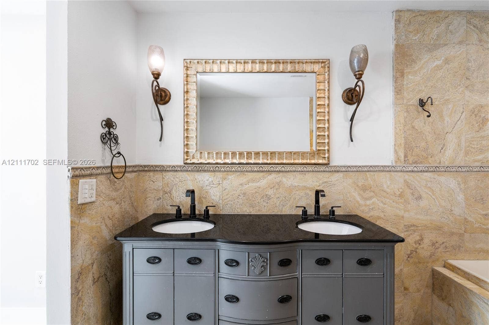 Dual-sink vanity with dark stone countertop, integrated undermount sinks, custom-finish cabinetry, ornate metallic wall sconces, and neutral stone tile wall surrounds