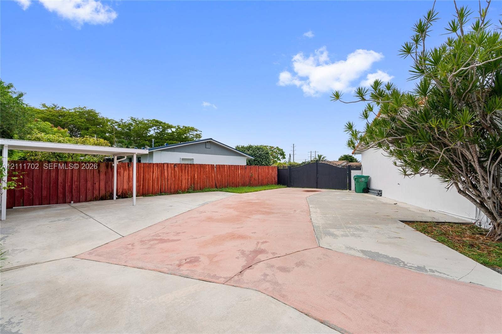 Concrete parking area featuring a covered carport, enclosed by a red wood-panel fence and a black metal entry gate