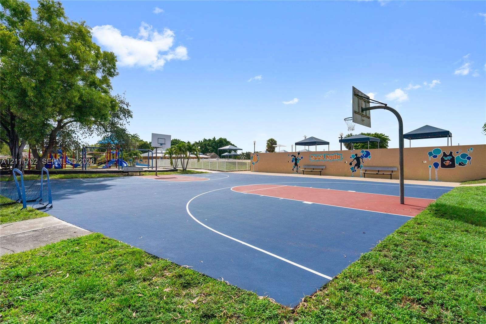 Outdoor multi-sport court featuring a paved basketball surface, metal backboards, perimeter bench seating, and shaded structures
