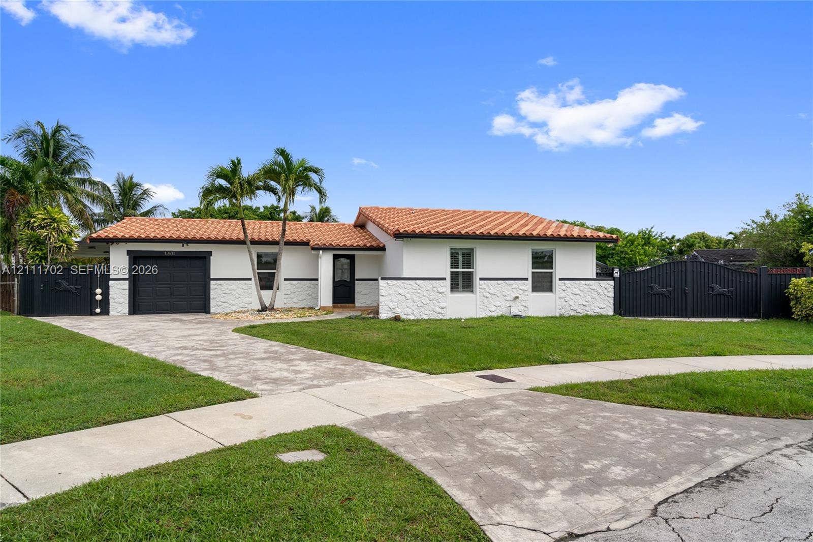 Single-story exterior featuring a red tile roof, white textured stone facade, attached garage with black door, and black perimeter fencing